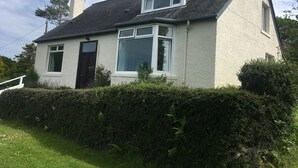 A two-story house with a sloped roof, white exterior walls, and a dark-colored door. - Detached house with stunning garden in picturesque coastal village SW Scotland (Kippford)