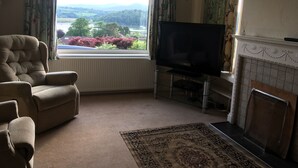 A living room with a sofa, a television on a stand, a fireplace, and a view of a landscape through a window. - Detached house with stunning garden in picturesque coastal village SW Scotland (Kippford)