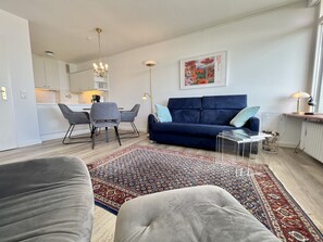 Un salon moderne avec un canapé bleu, des fauteuils gris, un tapis à motifs et un coin repas comprenant une table et des chaises.