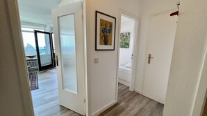 Un pasillo con piso de madera, paredes blancas y una puerta que da a una habitación con cama.