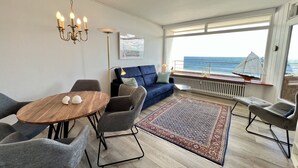 Una sala de estar moderna con un sofá azul, una mesa de comedor de madera y sillas grises. Se ve una ventana grande con vista al mar.