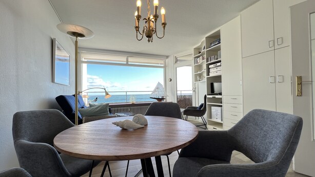 Un comedor con una mesa redonda de madera, sillas grises tapizadas, una araña, una estantería y vista al mar a través de un ventanal grande.