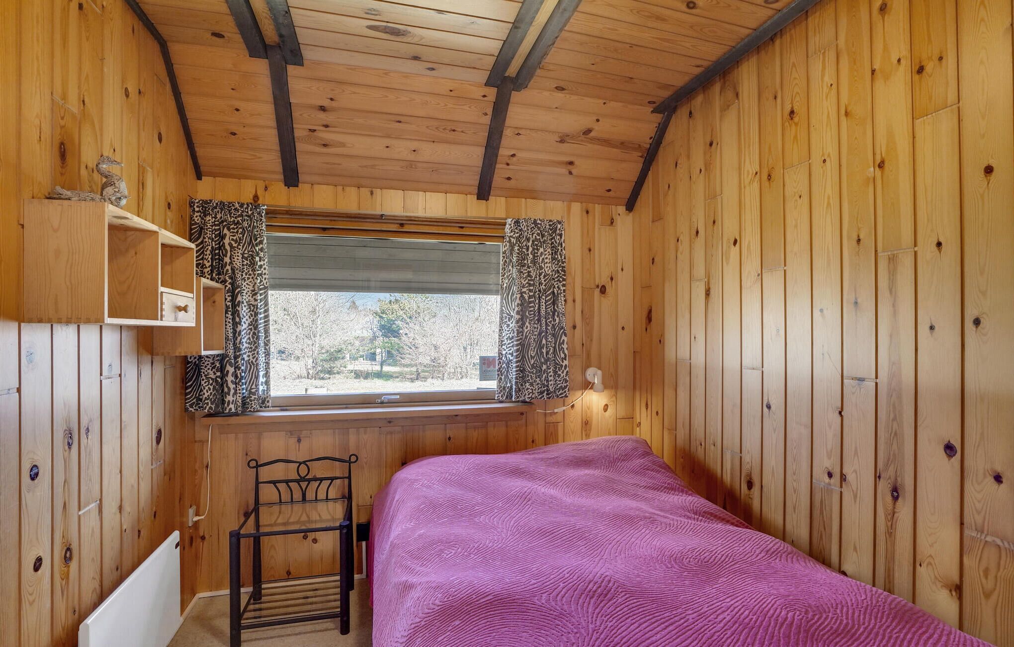 Una habitación de cabaña de madera con una cama, una ventana con cortinas estampadas, un estante y una silla.