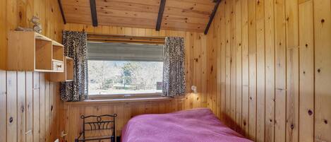 Una habitación de cabaña de madera con una cama, una ventana con cortinas estampadas, un estante y una silla.