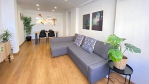 Un salon moderne avec un canapé d’angle gris, un coin repas comprenant une table et des chaises, et une grande plante en pot.