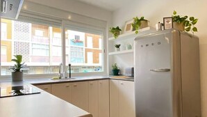 A modern kitchen with white cabinets, a built-in oven, and a refrigerator. There are potted plants on the windowsill and a view of buildings outside. - Project Montalvão, Setúbal (Setúbal)