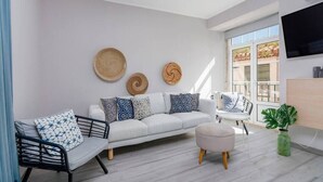 Un salon moderne avec un canapé blanc, des coussins à motifs, une chaise en métal noir, un pouf rond et une grande fenêtre laissant entrer la lumière naturelle.
