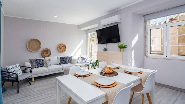 Un salon moderne avec un canapé blanc, une table à manger dressée avec des assiettes et des verres, et un téléviseur à écran plat fixé au mur.
