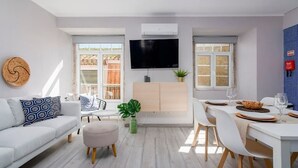 Un salon moderne comprenant un canapé, un pouf, une table à manger, des chaises, un téléviseur et une plante.