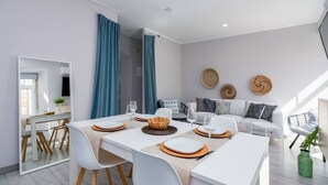 Un espace repas moderne avec une table blanche dressée pour quatre personnes, un canapé agrémenté de coussins à motifs et un grand miroir qui reflète la pièce.
