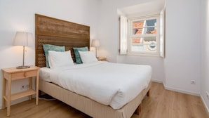 Een slaapkamer met een houten hoofdbord, twee nachtkastjes met lampen, een raam met uitzicht op een stadsbeeld, en een bed met witte beddengoed.