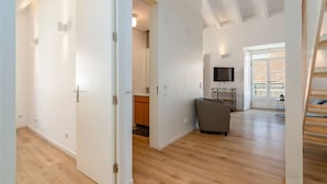 Een ruime woonkamer met houten vloer, een grijze bank, een aan de muur bevestigde televisie en een trap naar een hoger gelegen verdieping.