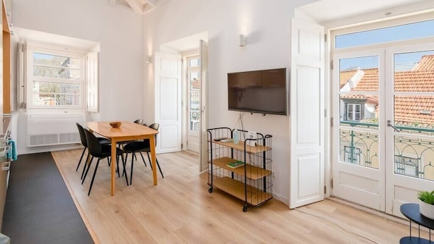 Een lichte kamer met een houten eettafel, stoelen, een televisie en een karretje met planken.