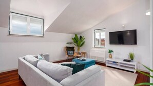 Ein modernes Wohnzimmer mit einem grauen Sofa, einem blauen Hocker, einem an der Wand befestigten Fernseher und einem großen Fenster mit Jalousien.