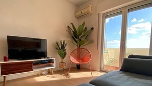 A modern living room with a flat-screen TV, a wooden entertainment unit, a red chair, a large potted plant, and a balcony with a view. - Setúbal Downtown SoHo (Setúbal)