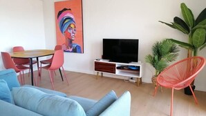 A modern living room with a blue sofa, a dining table with red chairs, a flat-screen TV on a white stand, and a large painting on the wall. - Setúbal Downtown SoHo (Setúbal)