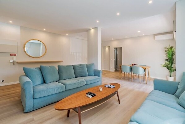 Een moderne woonkamer met een blauwe bank, een houten salontafel en een eethoek met een tafel en stoelen.