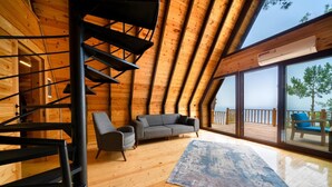 Intérieur moderne d’une cabane en rondins, avec un escalier en bois, un canapé gris et une grande fenêtre donnant sur une terrasse.