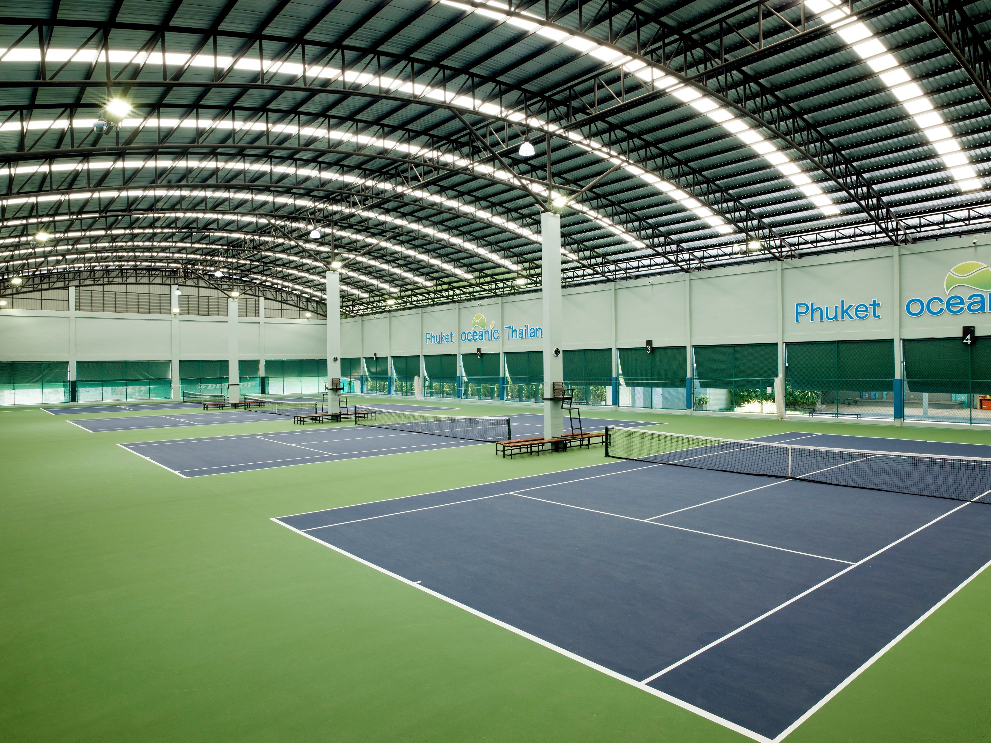 Pista de tenis interior con varias canchas, techo alto con vigas metálicas y una amplia pared verde con señalización.