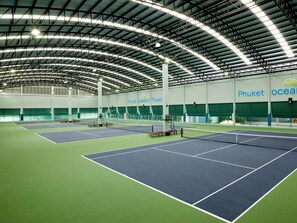 Pista de tenis interior con varias canchas, techo alto con vigas metálicas y una amplia pared verde con señalización.