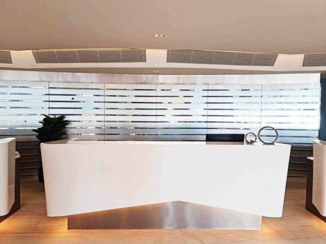 A modern reception desk with a sleek design, a large window with blinds, and a plant to the left.