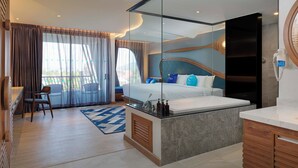 Una habitación de hotel moderna con una cama grande, un área de descanso con una silla, un escritorio pequeño y un baño con espejo y lavamanos.