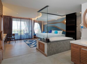 A modern hotel room with a large bed, a sitting area with a chair, a small desk, and a bathroom with a mirror and sink. - The Oceanic Sportel (Ko Kaeo)