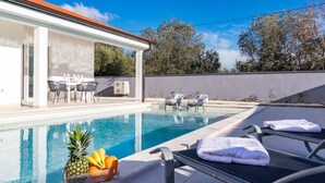 A swimming pool with lounge chairs and a table with chairs, a fruit basket, and a covered outdoor area with a table and chairs.