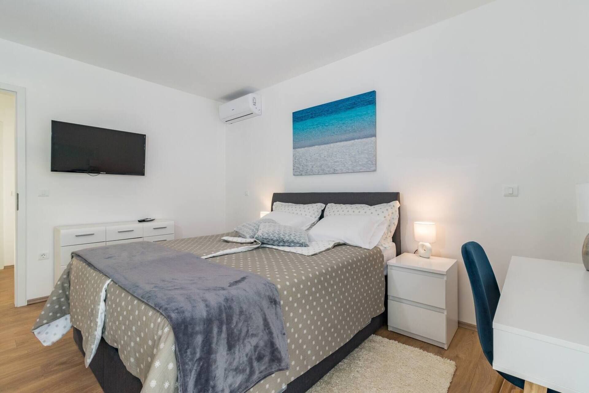Camera da letto moderna con un letto, comodini, una scrivania, una sedia, una televisione e un quadro a tema spiaggia.