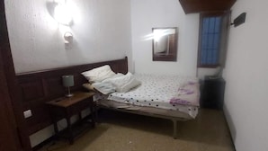 A small, dimly lit room with a single bed, a nightstand, a wall-mounted mirror, and a window with bars.