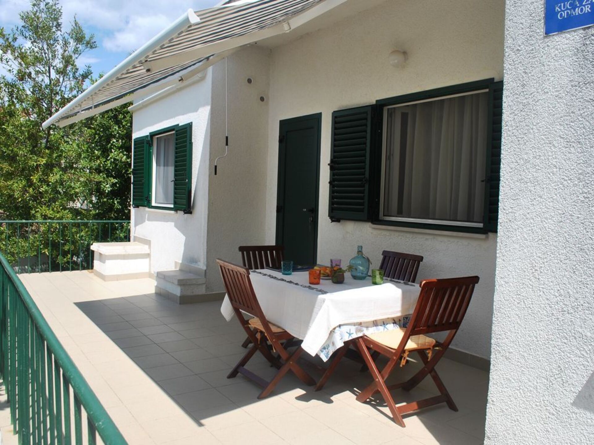 Une terrasse avec une table et des chaises, un bâtiment blanc avec des volets verts, et une enseigne sur laquelle on peut lire « KUC A ZADOMOR ».