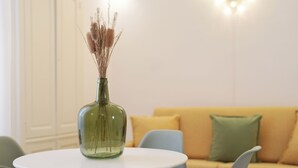 Una mesa redonda con un jarrón verde que contiene plantas secas, un sofá amarillo con cojines verdes, y una silla azul claro en un salón moderno.