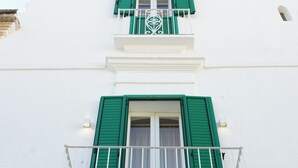 Un edificio de paredes blancas y contraventanas verdes, con un balcón y barandilla blanca.