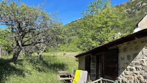 Un bâtiment rustique en pierre, avec un banc en bois et une table à l’extérieur, entouré d’une végétation luxuriante et d’arbres.