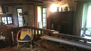 Une pièce rustique avec un banc en bois, une chaise et une table. On y trouve des fenêtres avec des rideaux, une étagère avec divers objets et un mur orné d’une horloge.