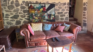 Un canapé en cuir marron agrémenté de plusieurs coussins, une petite table en bois avec deux télécommandes, et un tapis coloré posé sur un sol carrelé.