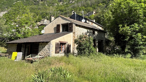 Une maison en pierre avec un toit en pente, entourée d’une végétation luxuriante et dominée par un paysage montagneux.