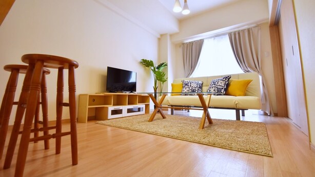 A living room with a sofa, TV, wooden stools, and a plant. - Chamomile Nishimachi 202 (Naha)