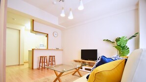 A modern living room with a glass coffee table, a sofa with yellow cushions, a TV, and a plant. - Chamomile Nishimachi 202 (Naha)