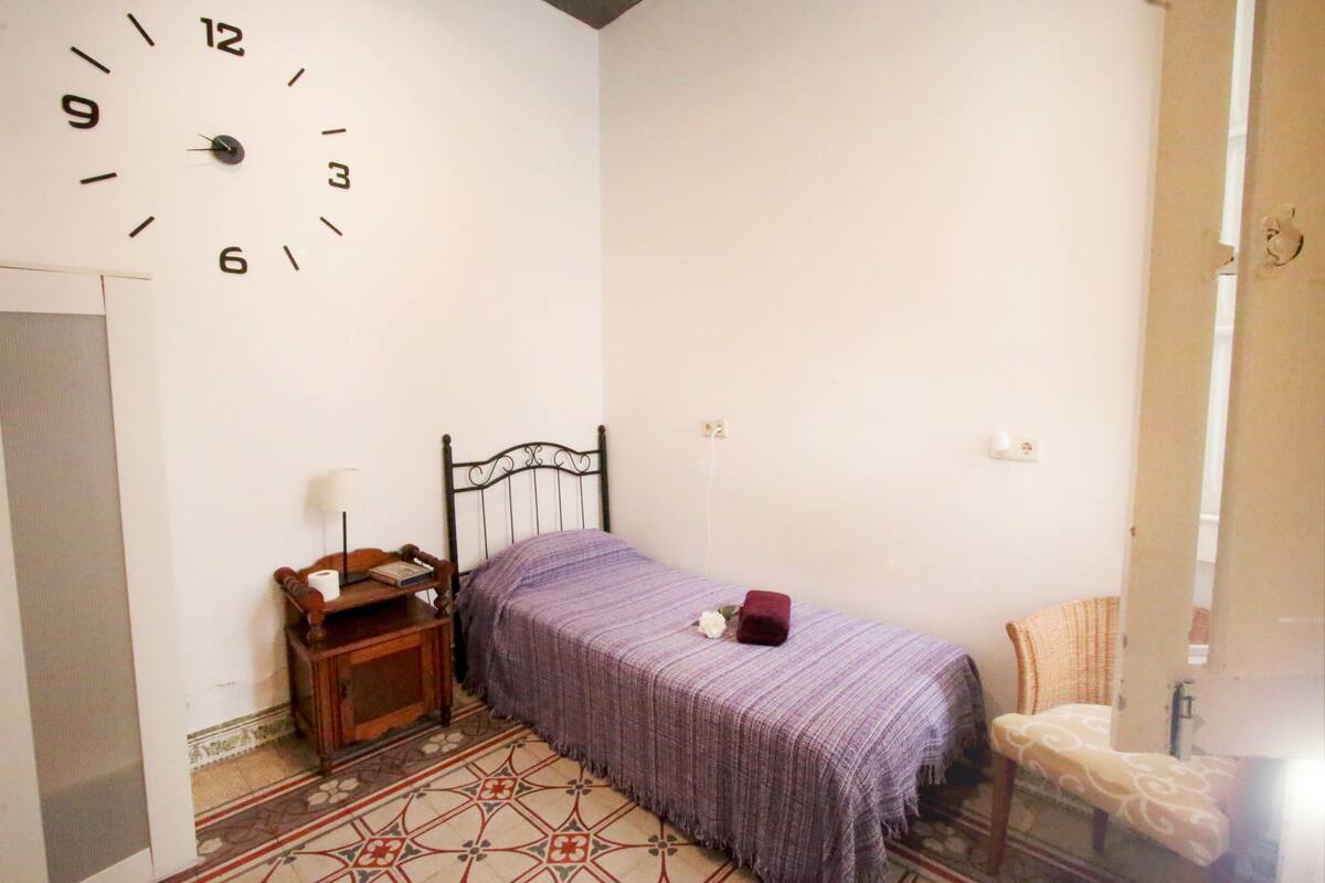 Una habitación pequeña y sencilla con cama, una silla, una mesita de noche y un reloj de pared.