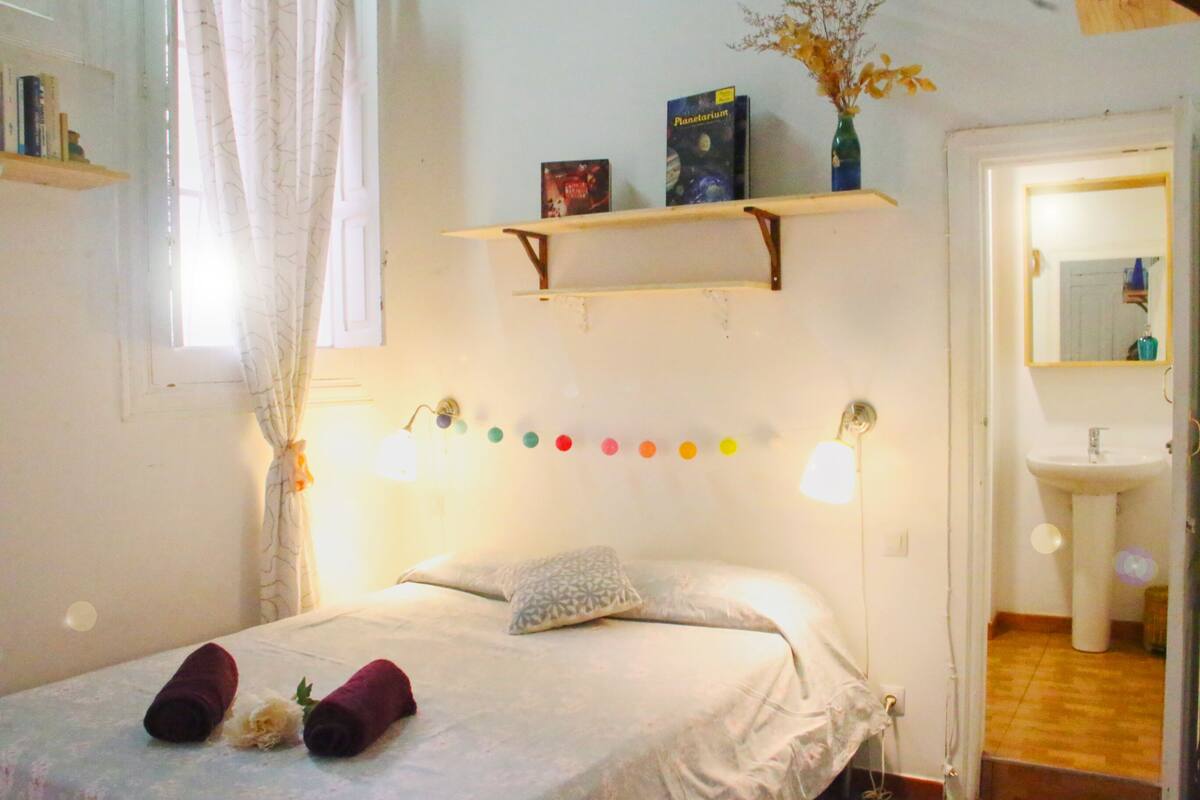 Un dormitorio con cama, almohadas, una estantería con libros, un espejo y un baño visible a través de una puerta abierta.
