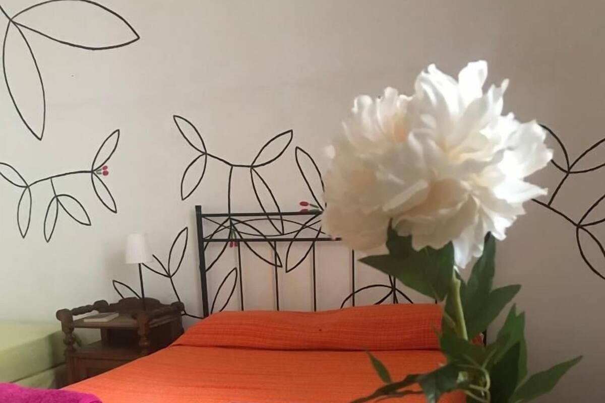 Habitación de hotel con una cama, una mesita de noche de madera, una mesa con una botella de vino y un florero con una flor blanca.