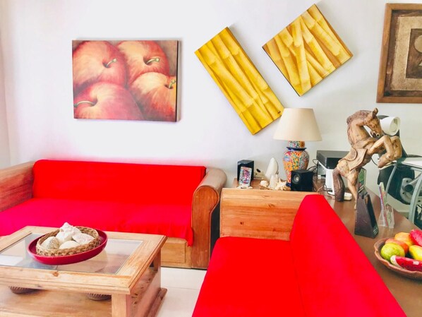 Una sala de estar con un sofá rojo, una mesa de centro de madera con un cesto de galletas y una pared decorada con dos obras enmarcadas.