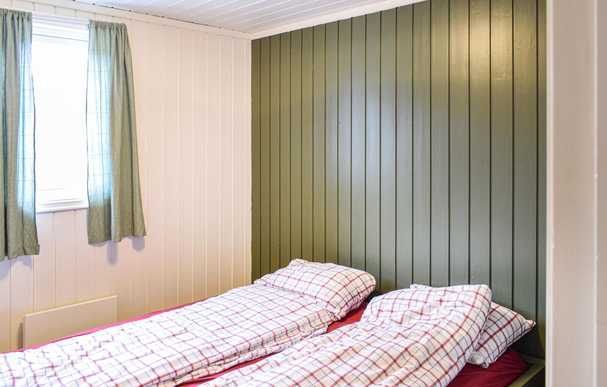 Un lit simple avec une couvre-lit à carreaux et des oreillers, une fenêtre avec des rideaux verts et un mur recouvert de panneaux de bois verticaux.