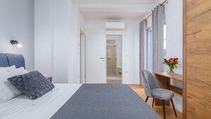 Quarto com cama, mesa de cabeceira, cadeira, escrivaninha, janela com cortinas e ar condicionado.