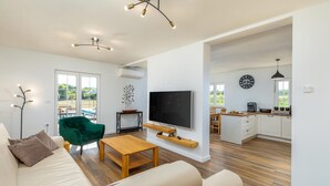 Ampla sala de estar com sofá, uma poltrona verde, mesa de centro de madeira e televisão de tela plana. Há uma área de cozinha com relógio e máquina de café.