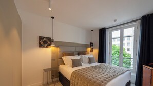 Une chambre moderne avec un grand lit, des tables de chevet, une tête de lit et une fenêtre donnant sur des immeubles et des arbres.