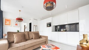 Ett modernt vardagsrum med en beige soffa, ett soffbord med böcker, en köksdel med vita skåp och en röd hänglampa.