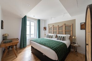 Ein Schlafzimmer mit Bett, Schreibtisch, Fenster mit Vorhängen, einem hölzernen Kopfteil und einer Tür.