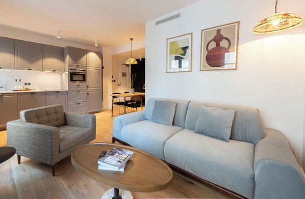 A modern living room with a grey sofa, a grey armchair, and a wooden coffee table. There is a kitchen in the background with grey cabinets and a dining area with a table and chairs. - Diodati, 4 stars, Elegente & Trendy at few meters away from the Champs-Elysées (París)